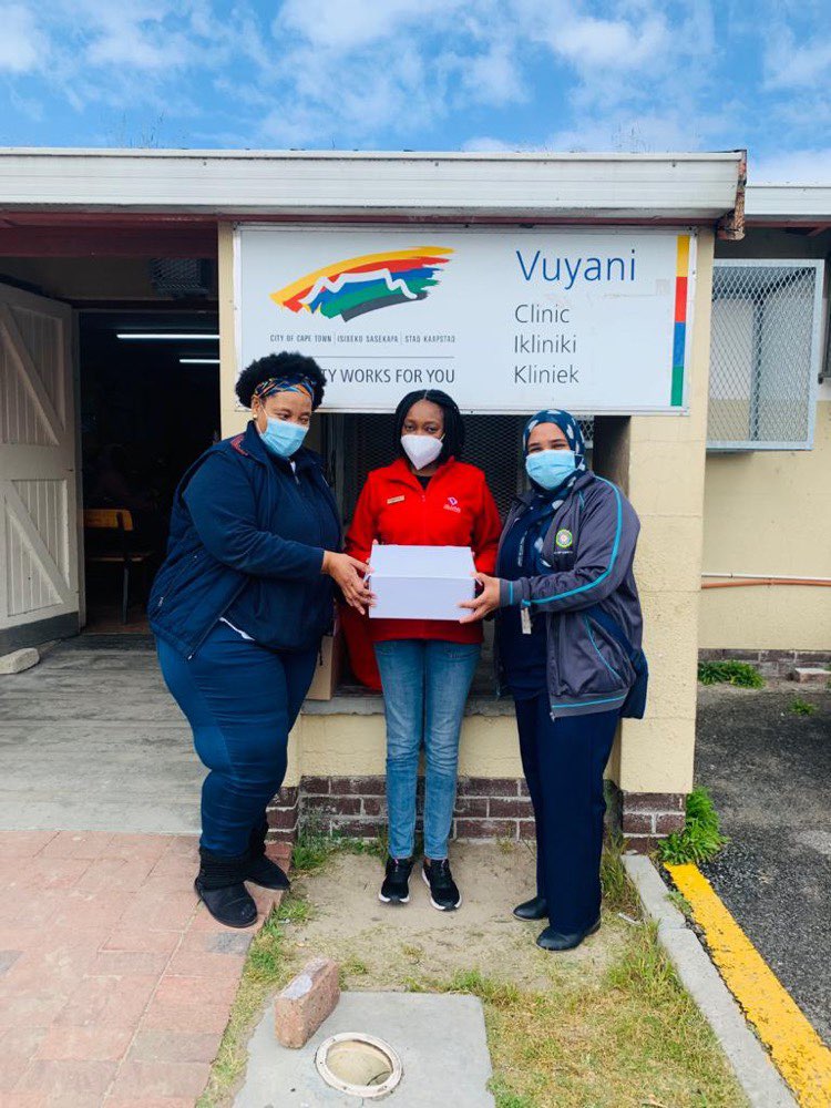 ASCENT team member with health facility workers holding a white box in front of the health facility "Vuyani" clinic. The three of them are wearing face masks.