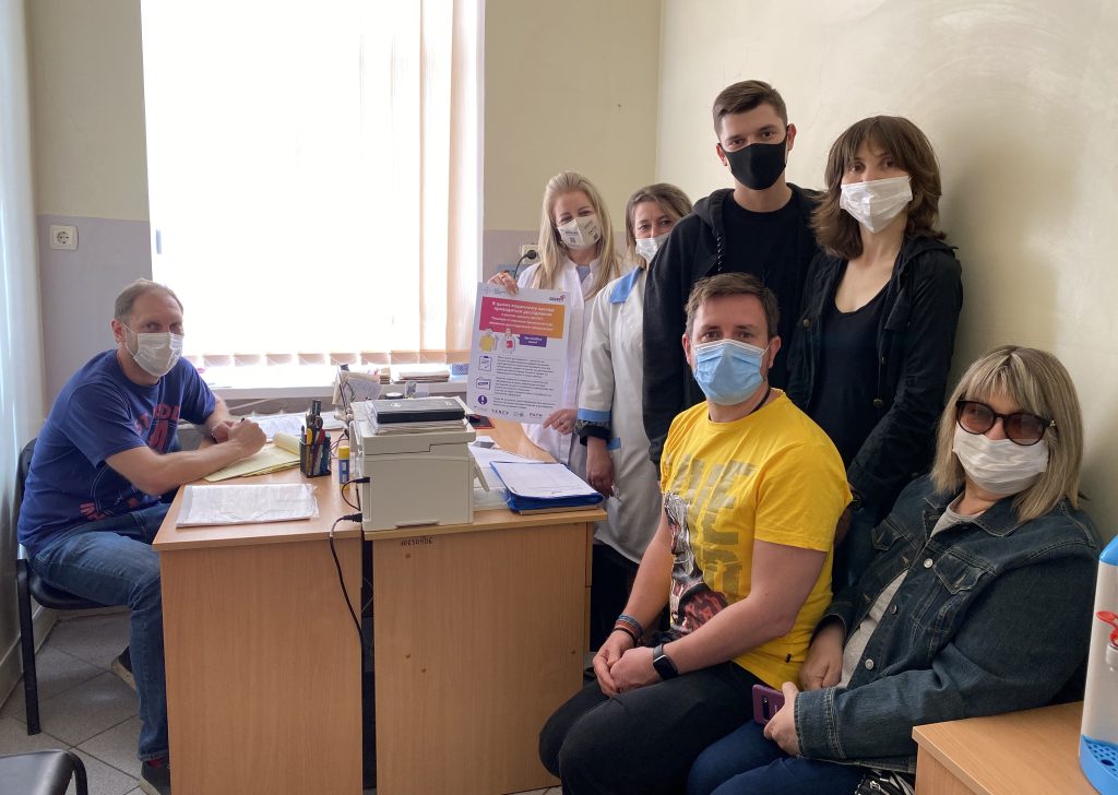 Seven people in a health facility office. All of them are wearing face masks. Two people are dressed with healthcare provider uniform.